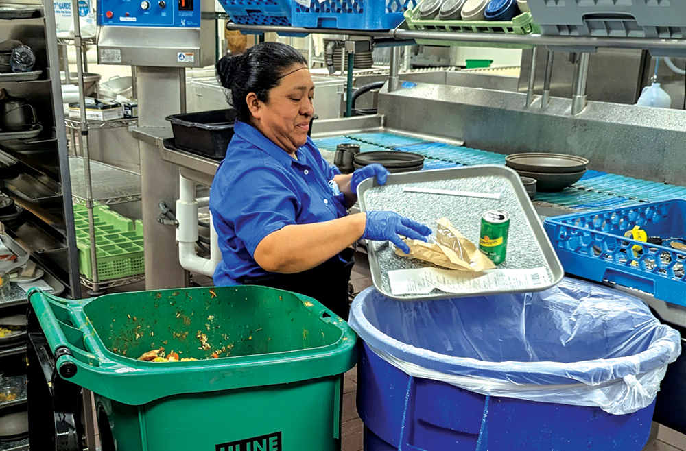 In a kitchen, a worker sorts items from a tray into waste bins.